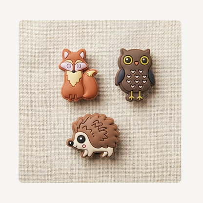 Three small animal-shaped clog charms arranged on a textured beige fabric background: a brown fox, a dark brown owl, and a hedgehog.