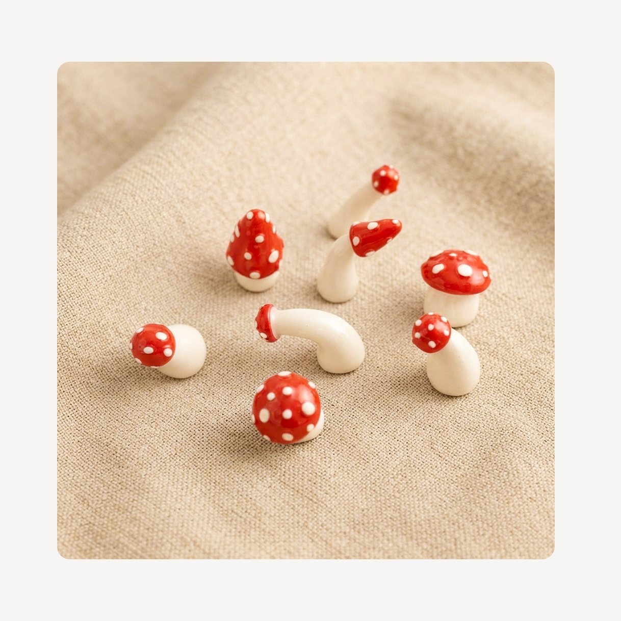 A set of small decorative mushroom clog charms with red caps and white spots, arranged on a beige textured fabric background.