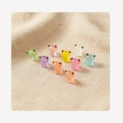 A group of small translucent frog-shaped  clog charms in pastel colors, including green, pink, yellow, white, purple, peach, and light blue, arranged on a textured beige fabric background.