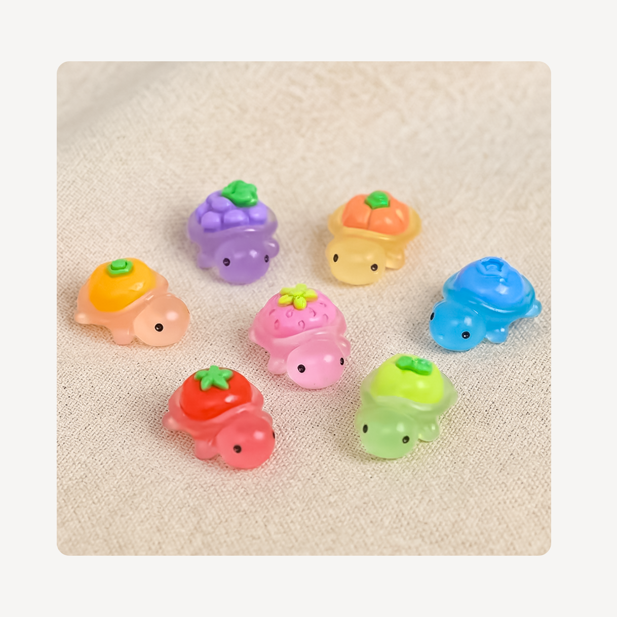 Seven small pastel turtle-shaped clog charms arranged on a light beige textured fabric background. Each turtle has a different body color and a fruit-themed shell with green tops, including orange, purple, peach, blue, pink, red, and green.
