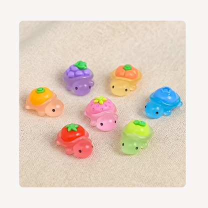 Seven small pastel turtle-shaped clog charms arranged on a light beige textured fabric background. Each turtle has a different body color and a fruit-themed shell with green tops, including orange, purple, peach, blue, pink, red, and green.