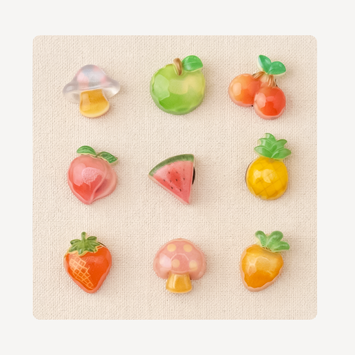 Nine small fruit-shaped clog charms arranged in a 3-by-3 grid on a textured beige square background. The designs include a mushroom, green apple, two cherries, peach, watermelon slice, pineapple, strawberry, pink mushroom, and orange-yellow fruit.