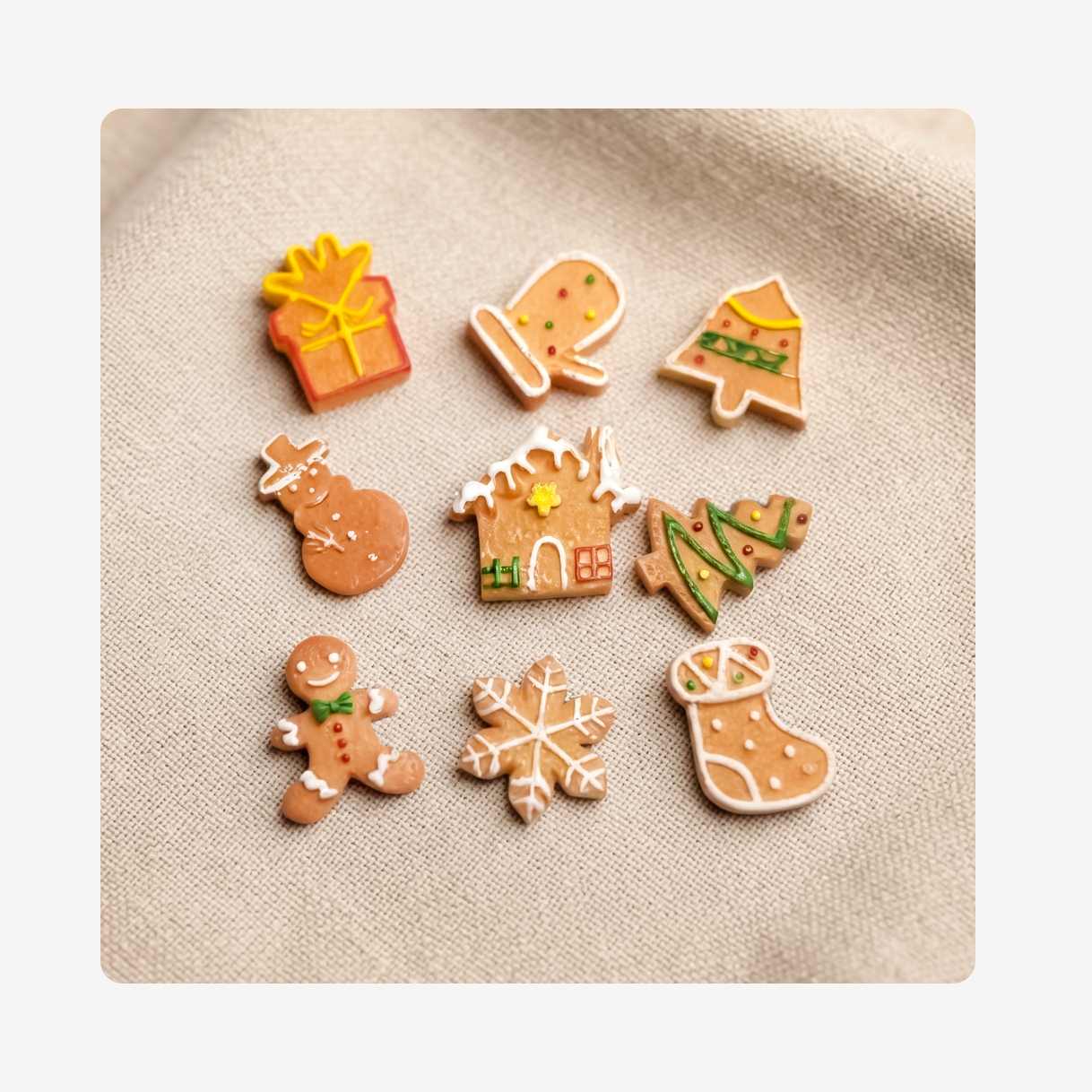 Nine small gingerbread-themed clog charms arranged on a beige fabric background, including a gift box, mitten, Christmas tree, snowman, house, Christmas tree, gingerbread man, snowflake, and stocking.