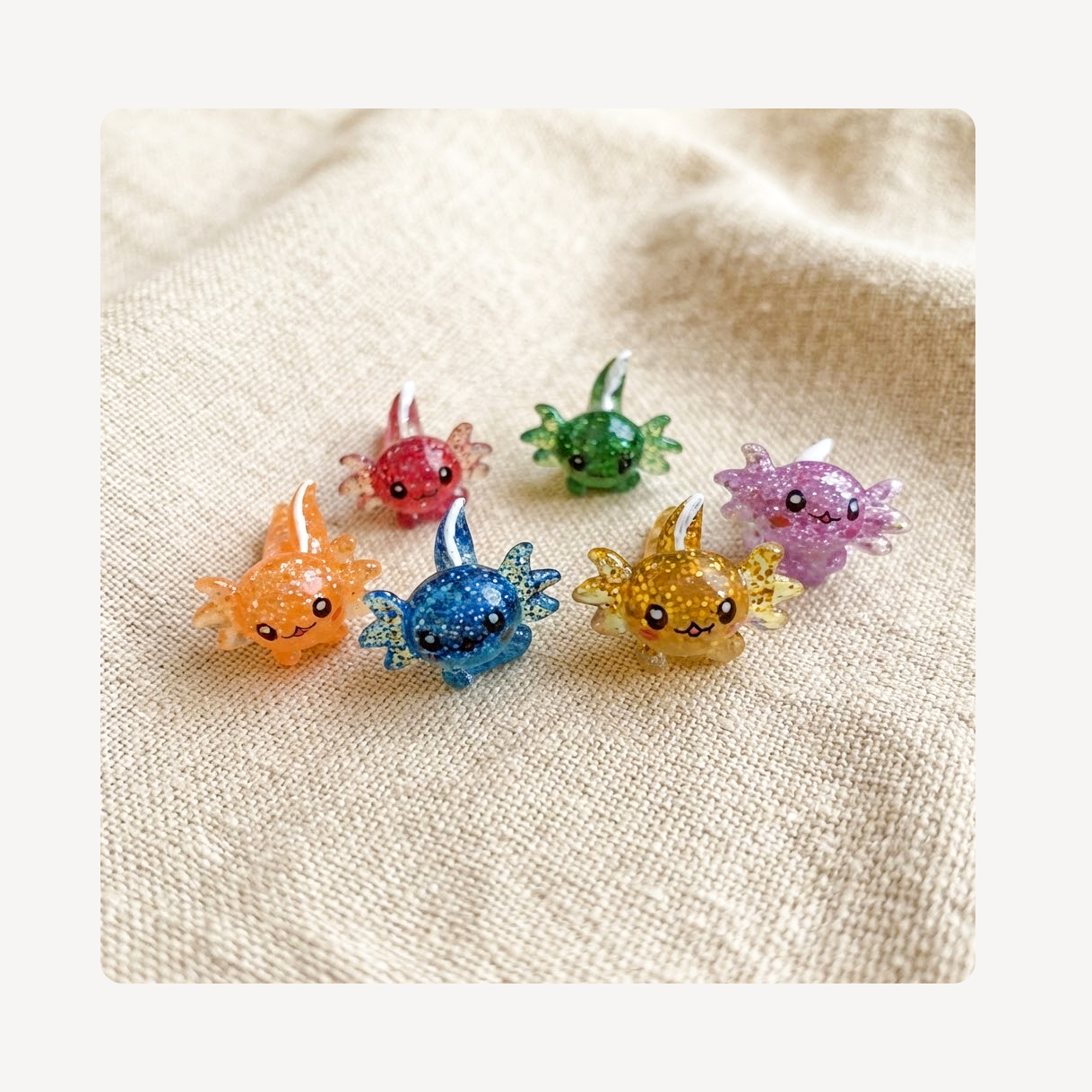 Six small glittery axolotl-shaped clog charms in assorted colors, including orange, blue, yellow, pink, green, and purple, arranged on a textured beige fabric background.