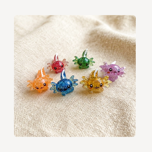 Six small glittery axolotl-shaped clog charms in assorted colors, including orange, blue, yellow, pink, green, and purple, arranged on a textured beige fabric background.