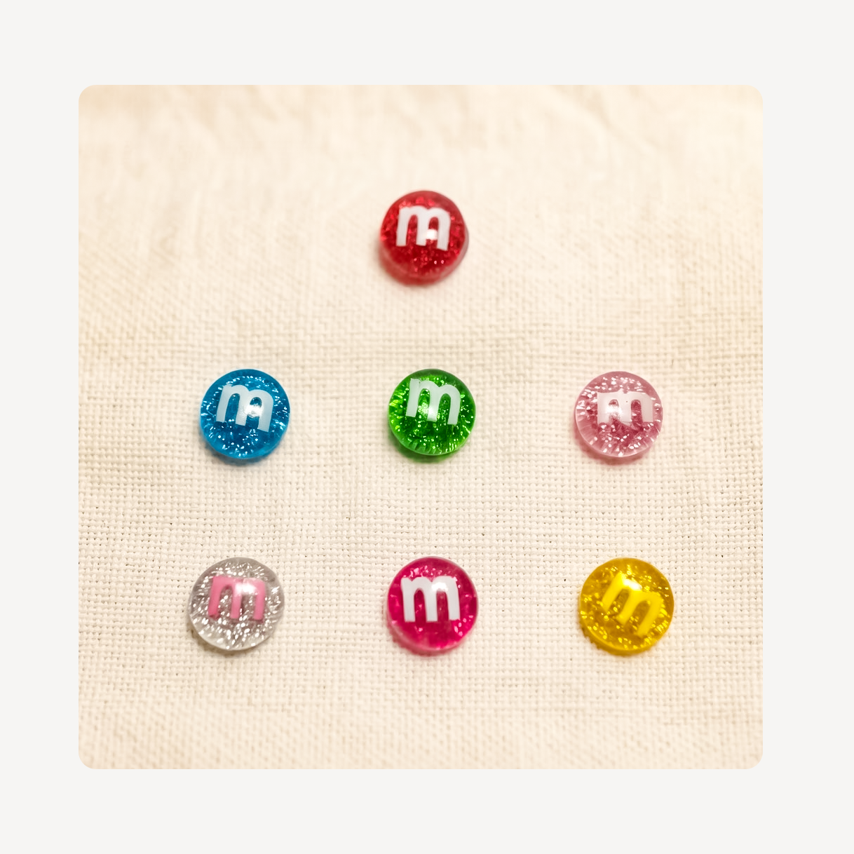 Six round glittery M&M's style clog charms with white 'm' logos, arranged on a textured cream fabric background.