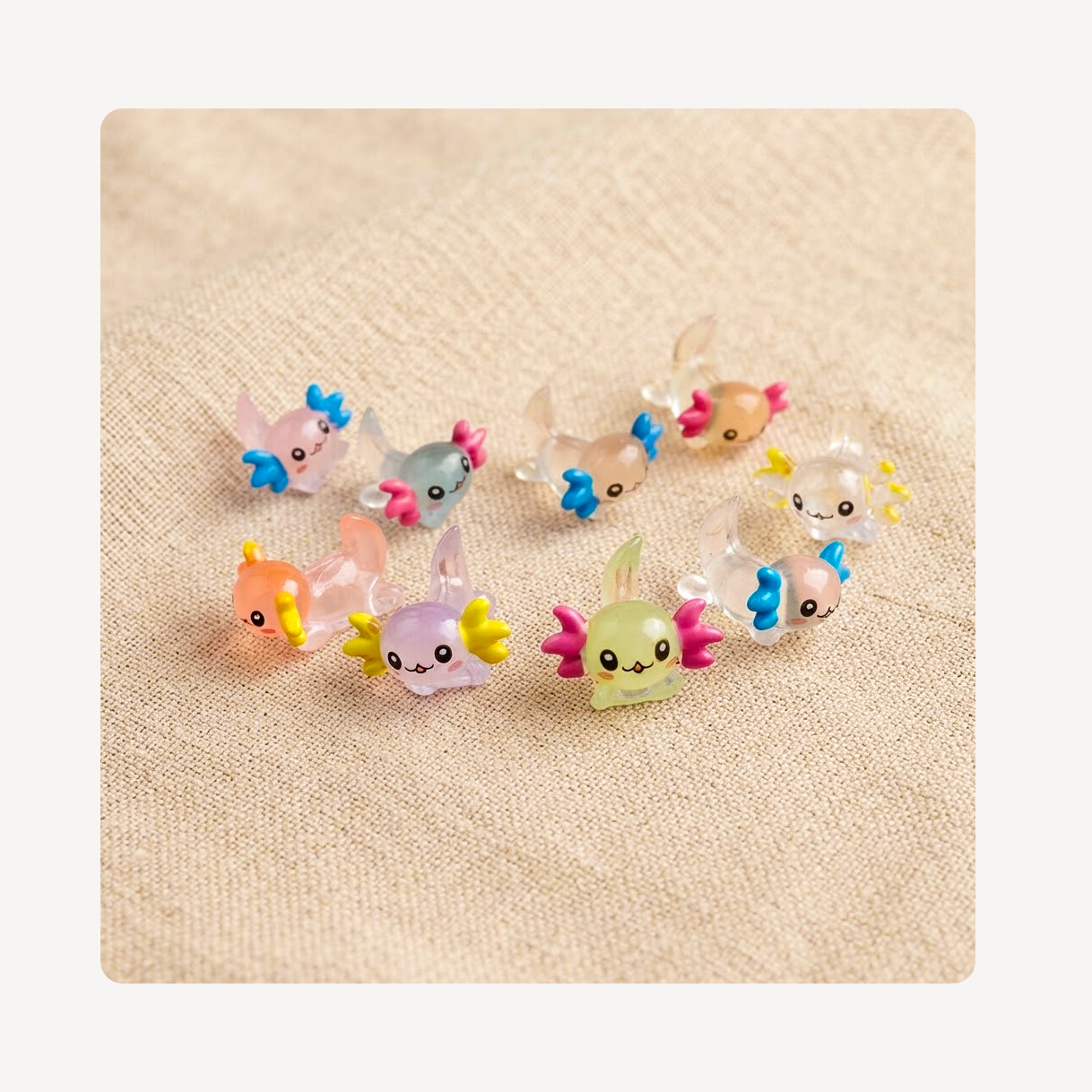 A set of nine small, colorful clog charms shaped like cute axoltols, arranged on a textured beige fabric background.