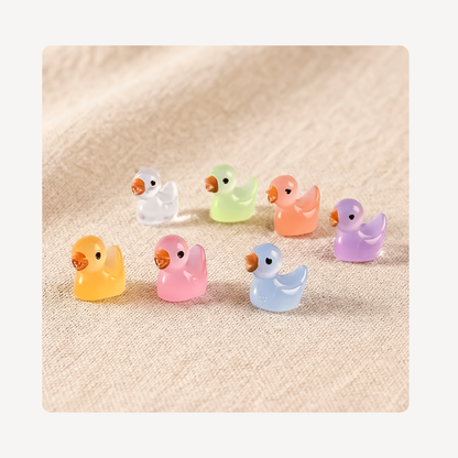 Seven small translucent duck-shaped clog charms in pastel colors including yellow, pink, light blue, orange, green, purple, and white, arranged on a beige textured fabric background.