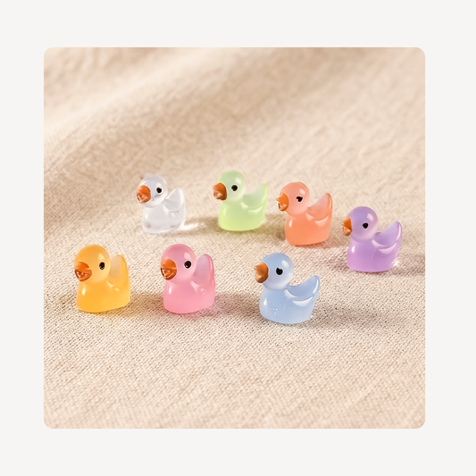 Seven small translucent duck-shaped clog charms in pastel colors including yellow, pink, light blue, orange, green, purple, and white, arranged on a beige textured fabric background.