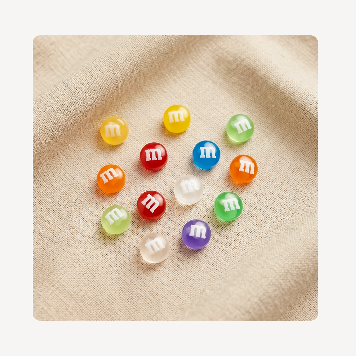 Ten colorful glossy candy-like clog charms with white lowercase 'm' logos, arranged on a beige fabric background.