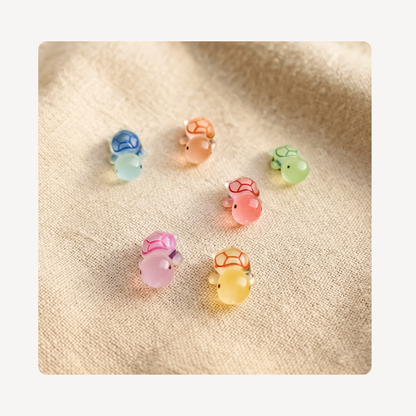 Six small translucent turtle-shaped clog charms in pastel colors, including blue, peach, green, pink, yellow, and purple, arranged on a beige textured fabric background.