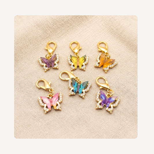 Six gold butterfly-shaped shoe charms with colorful enamel fills and clear rhinestone accents, arranged on a beige textured fabric background.