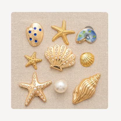 Nine seashell and starfish-shaped clog charms arranged on a square beige fabric background. The set includes gold-tone starfish and shell charms with faux pearls, blue and green stone accents, and one larger gold conch shell.