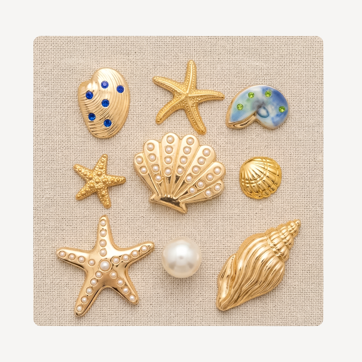 Nine seashell and starfish-shaped clog charms arranged on a square beige fabric background. The set includes gold-tone starfish and shell charms with faux pearls, blue and green stone accents, and one larger gold conch shell.