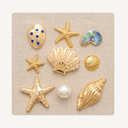 Nine seashell and starfish-shaped clog charms arranged on a square beige fabric background. The set includes gold-tone starfish and shell charms with faux pearls, blue and green stone accents, and one larger gold conch shell.