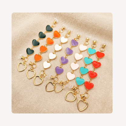 Seven pairs of gold heart clog charm chains arranged in a fan shape on a beige textured fabric background. Each pair features a combination of glossy heart charms in various colors (green, orange, white, purple, pink, teal, red) and open heart charms.