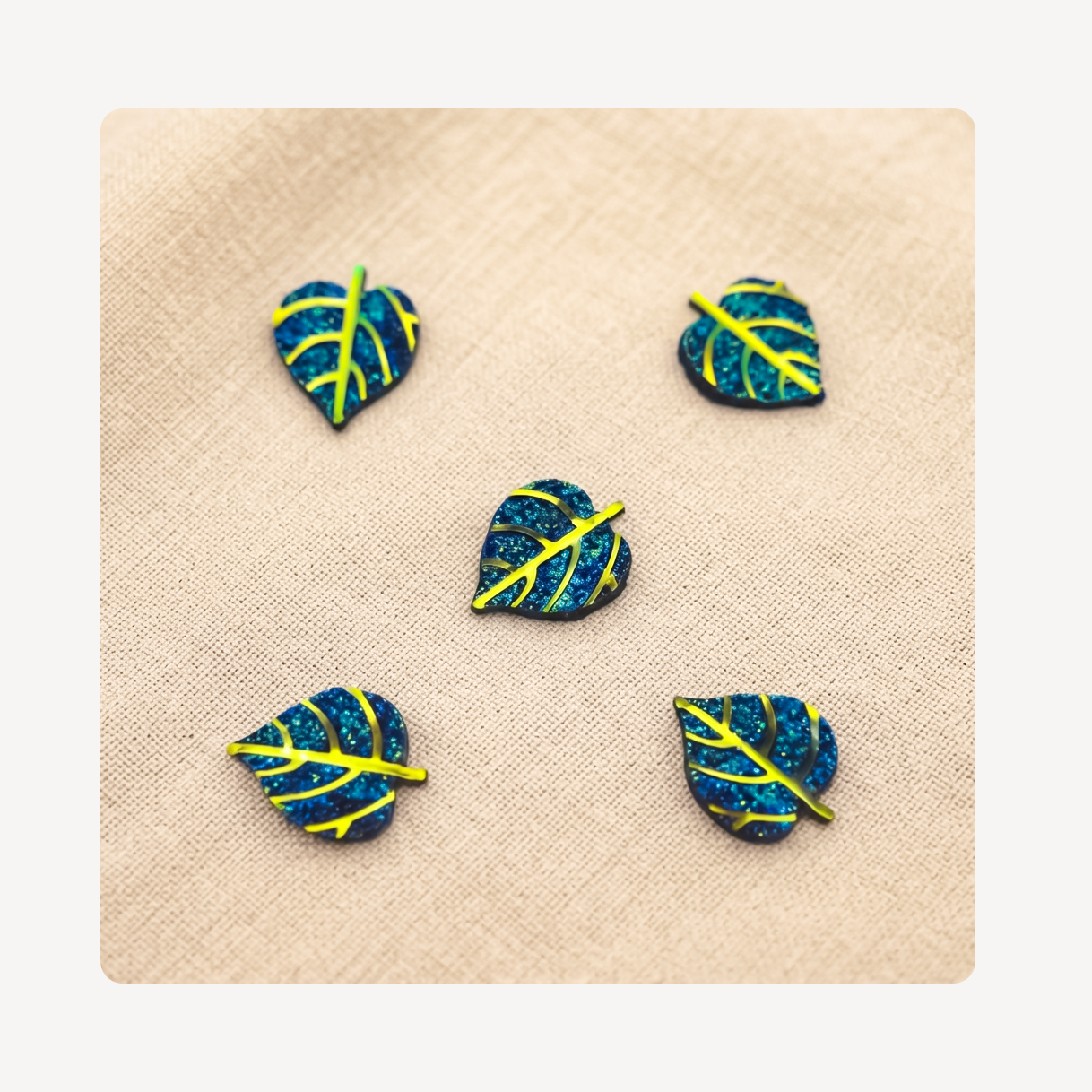 Five small leaf-shaped clog charms with a blue glitter finish and bright yellow vein details, arranged on a beige textured fabric background.
