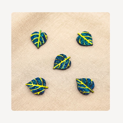 Five small leaf-shaped clog charms with a blue glitter finish and bright yellow vein details, arranged on a beige textured fabric background.