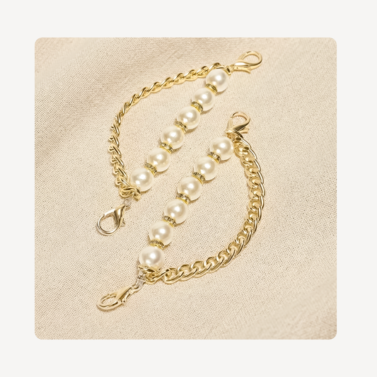 A pair of gold shoe chains with white pearl beads and lobster clasp connectors, displayed on a textured beige fabric background.