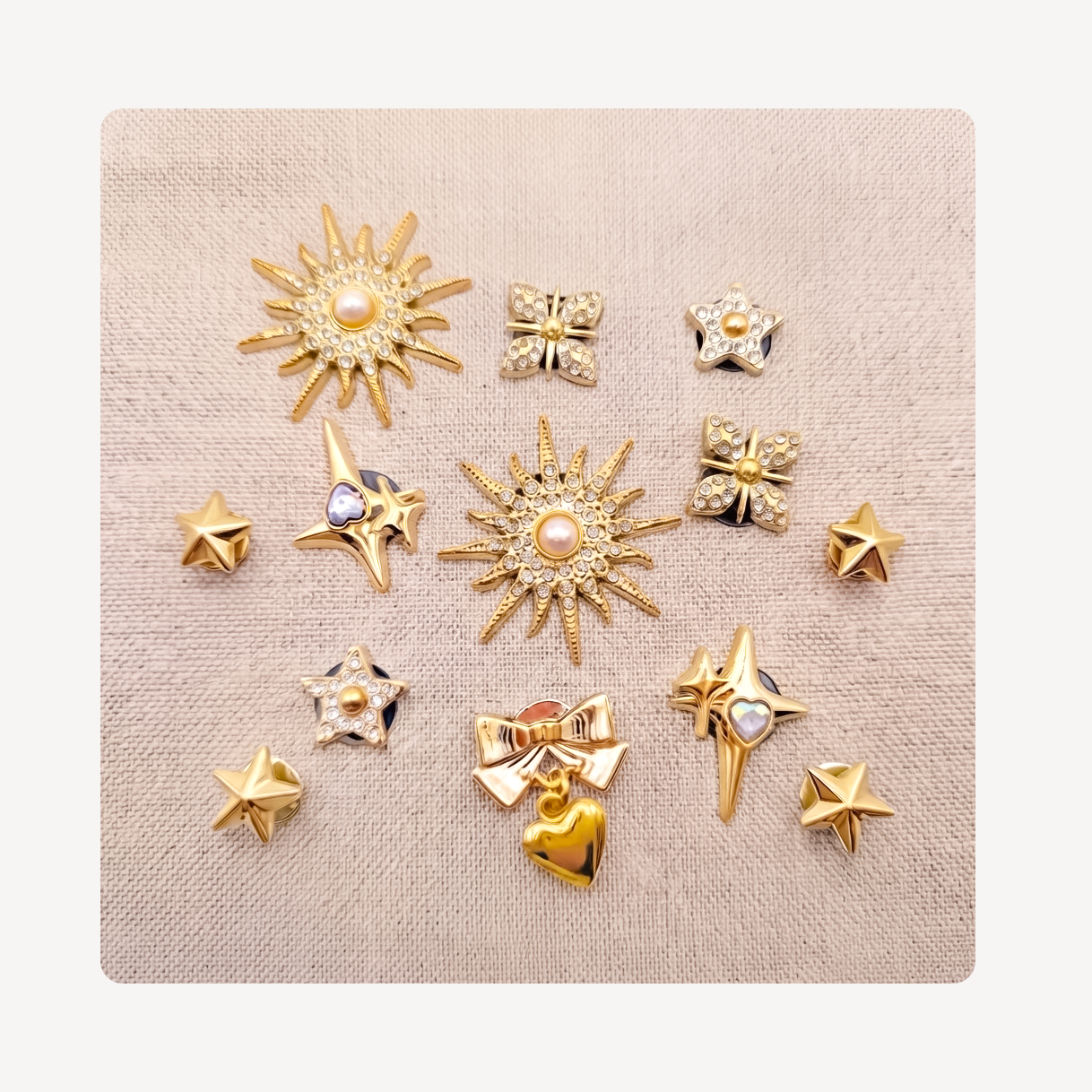 A set of 13 gold-toned clog charms arranged on a textured beige fabric background. The collection features various celestial and bow motifs, including sunbursts, stars, bows, and hearts, with some adorned with clear rhinestones and pearl-like center gems.