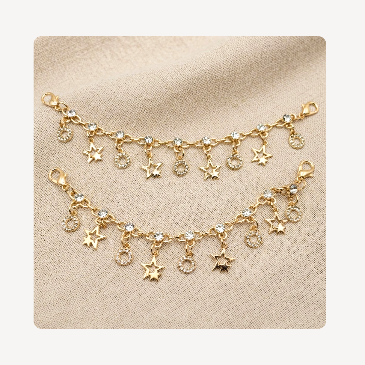 Gold-toned shoe chains featuring dangling star and crescent moon charms, some embellished with clear rhinestones, displayed on a textured beige fabric background.