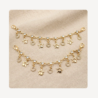 Gold-toned shoe chains featuring dangling star and crescent moon charms, some embellished with clear rhinestones, displayed on a textured beige fabric background.