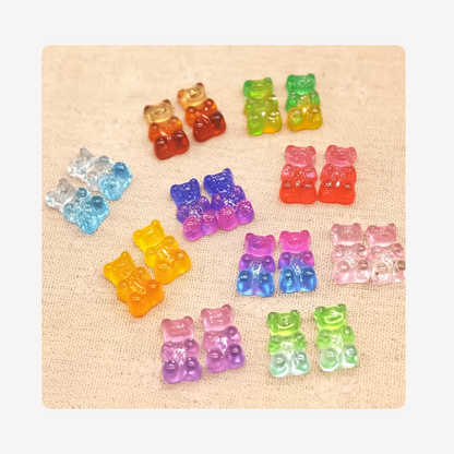 Twelve pairs of colorful translucent gummy bear clog charms arranged on a beige textured fabric background.