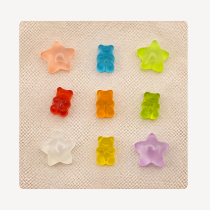Nine small translucent gummy candy clog charms arranged in a 3-by-3 grid on a textured beige square background. The candies include peach, blue, lime green, red, orange, yellow-green, clear/white, yellow, and lavender, with two star-shaped and seven bear-shaped pieces.