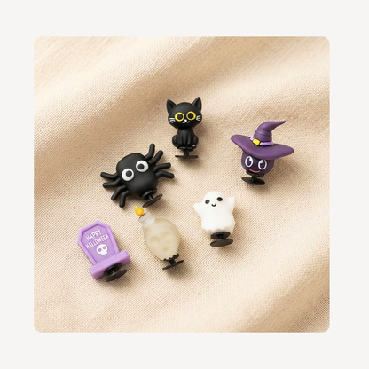 Five small Halloween-themed clog charms arranged on a beige fabric background: a black cat with yellow eyes, a black spider, a purple witch hat character, a white ghost, and a purple tombstone with 'Happy Halloween' text.