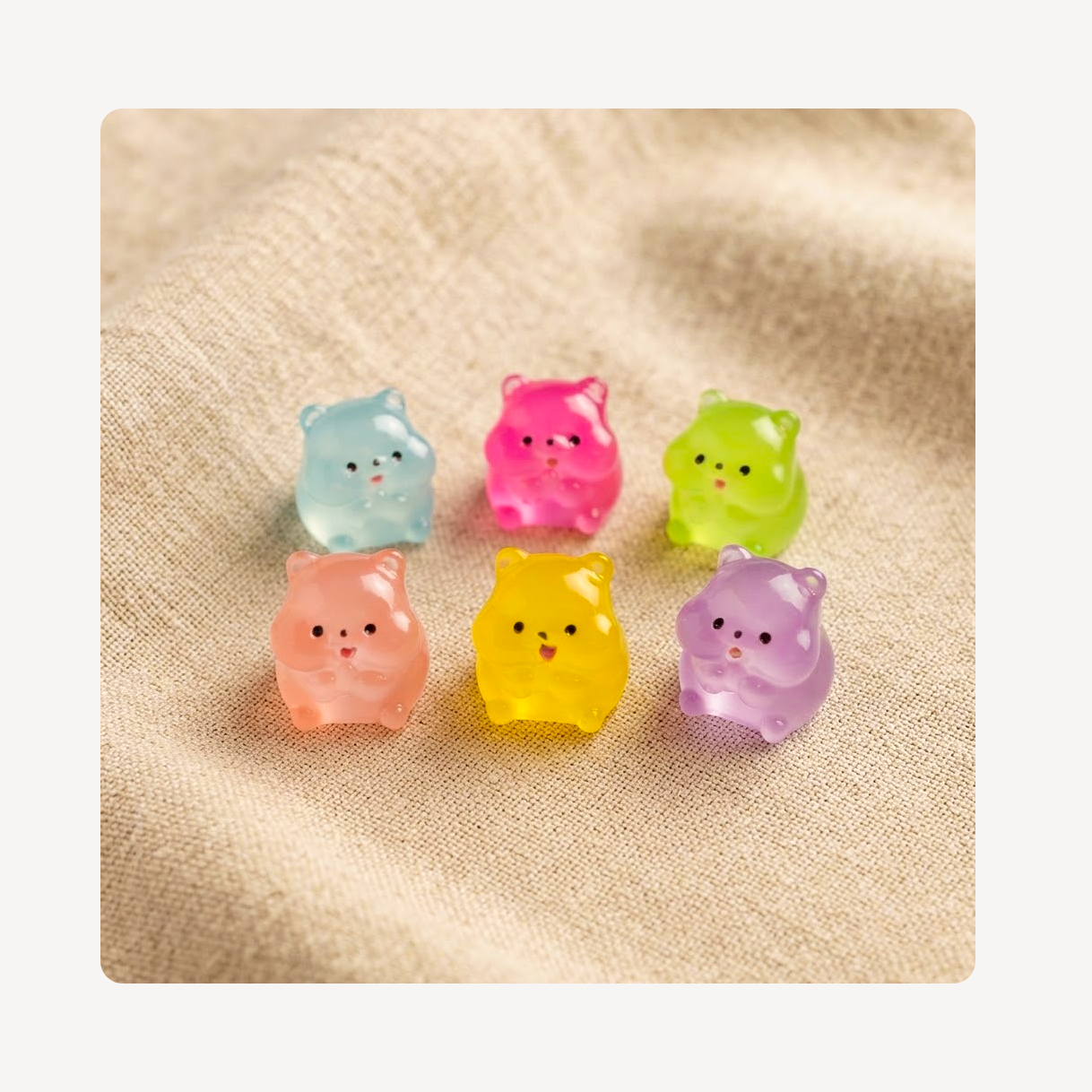 Six small translucent hamster-shaped clog charms in pastel colors, arranged on a beige textured fabric background.