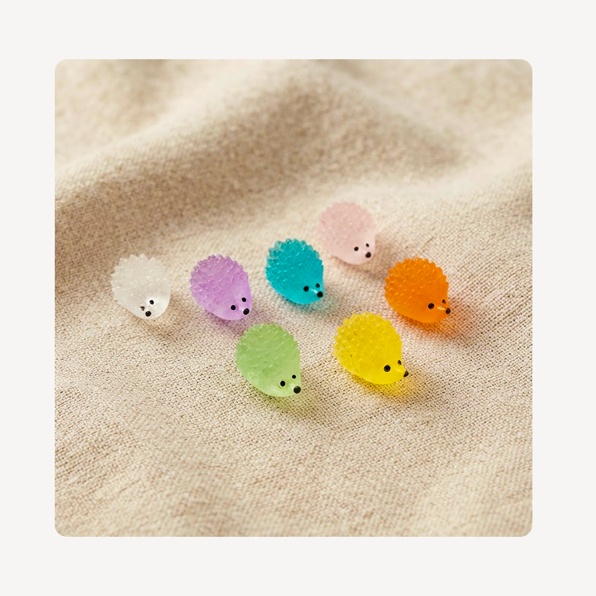 Six small hedgehog-shaped clog charms in pastel colors including white, purple, teal, pink, orange, green, and yellow, arranged on a soft beige textured fabric background.