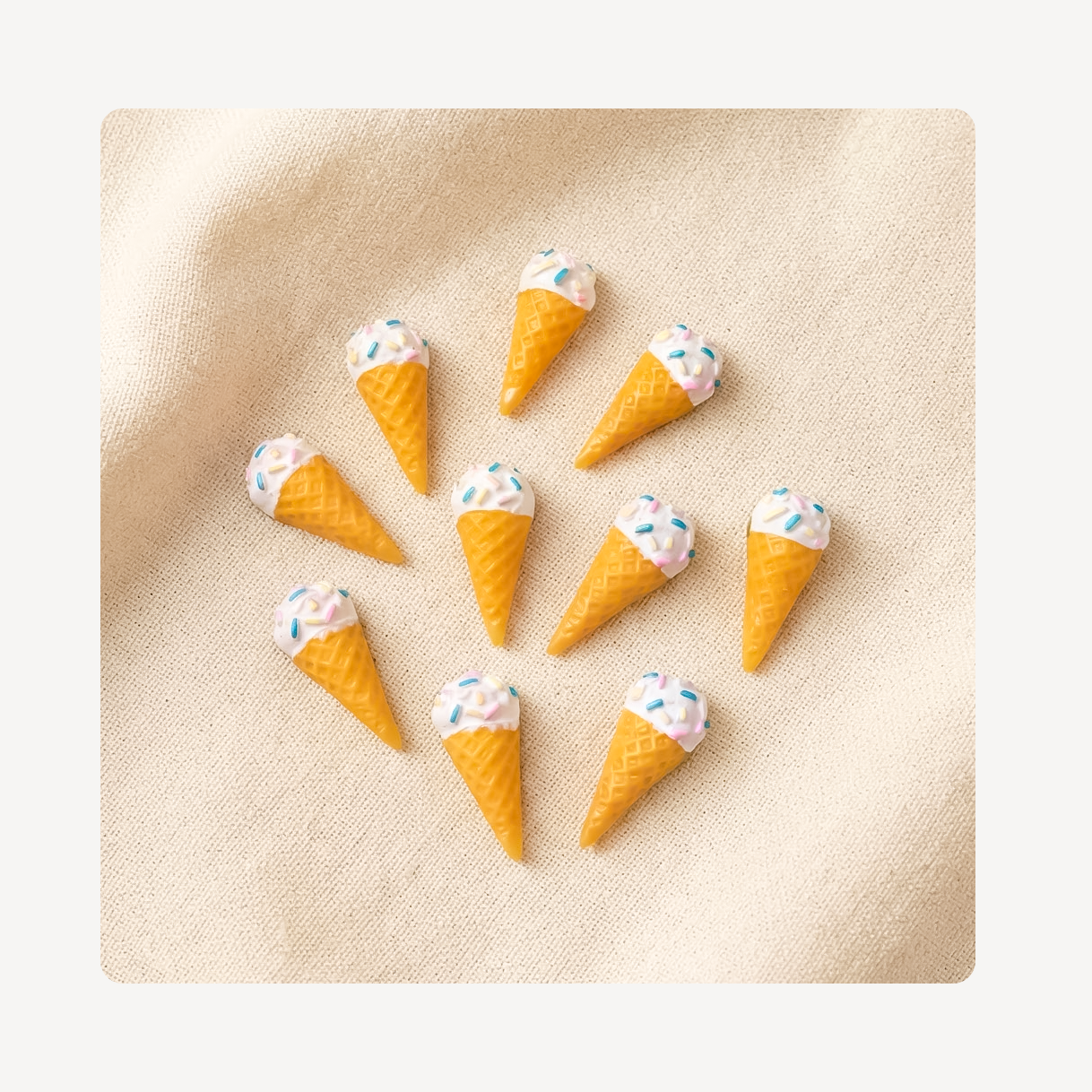 Nine miniature ice cream cone clog charms with waffle cones, white scoops, and pastel sprinkles, arranged on a textured beige fabric background.
