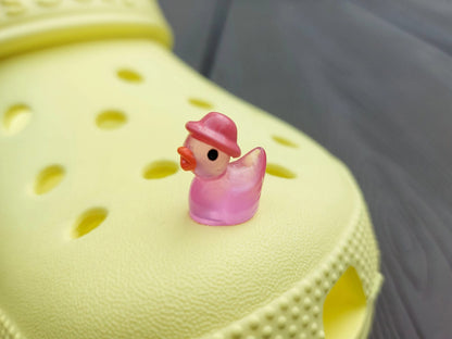 Glow in the dark duck charms wearing bucket hats on a yellow shoes shoe. These colorful charms are available in pink, white, blue, green, yellow, turquoise, peach and purple. The hats all have different colors.
