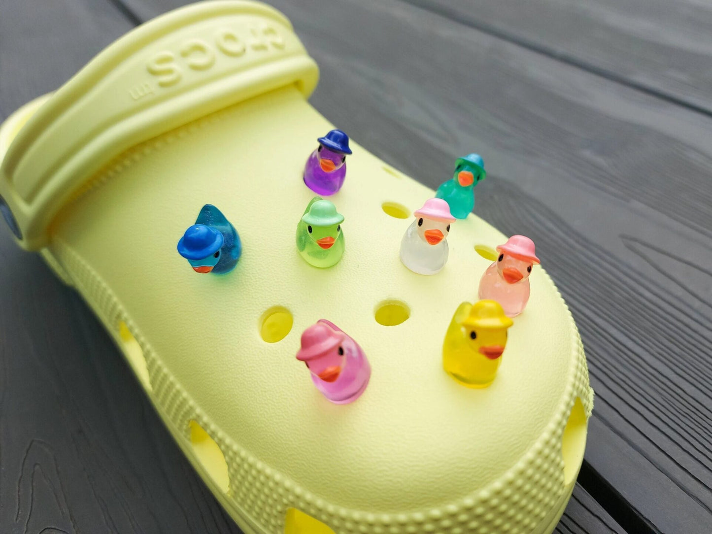Glow in the dark duck charms wearing bucket hats on a yellow shoes shoe. These colorful charms are available in pink, white, blue, green, yellow, turquoise, peach and purple. The hats all have different colors.