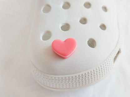 Yellow shoes adorned with pastel-colored macaron heart charms, placed among vibrant pink flowers. The charms add a playful and loving touch to the shoes.