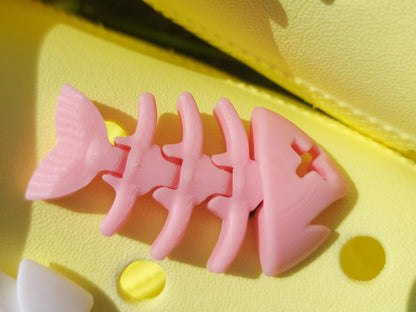 Yellow shoes adorned with colorful 3D-printed articulated fish bone charms in white, pink, black, teal, and yellow, displayed on a grassy background.