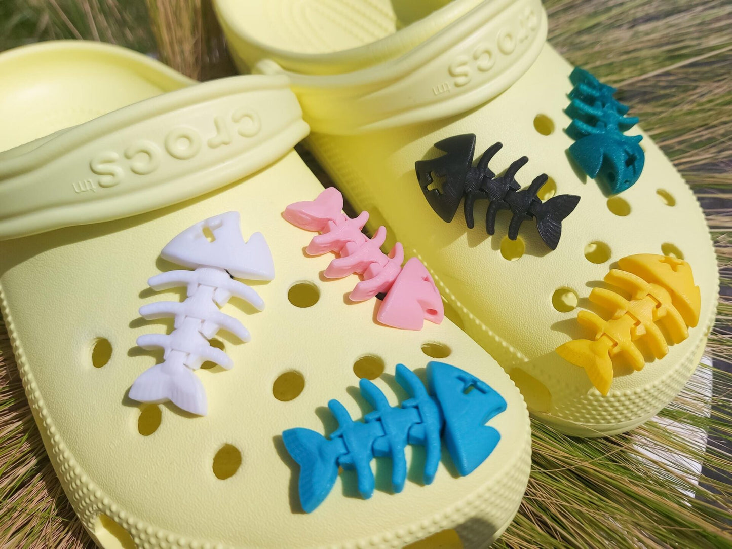 Yellow shoes adorned with colorful 3D-printed articulated fish bone charms in white, pink, black, teal, and yellow, displayed on a grassy background.