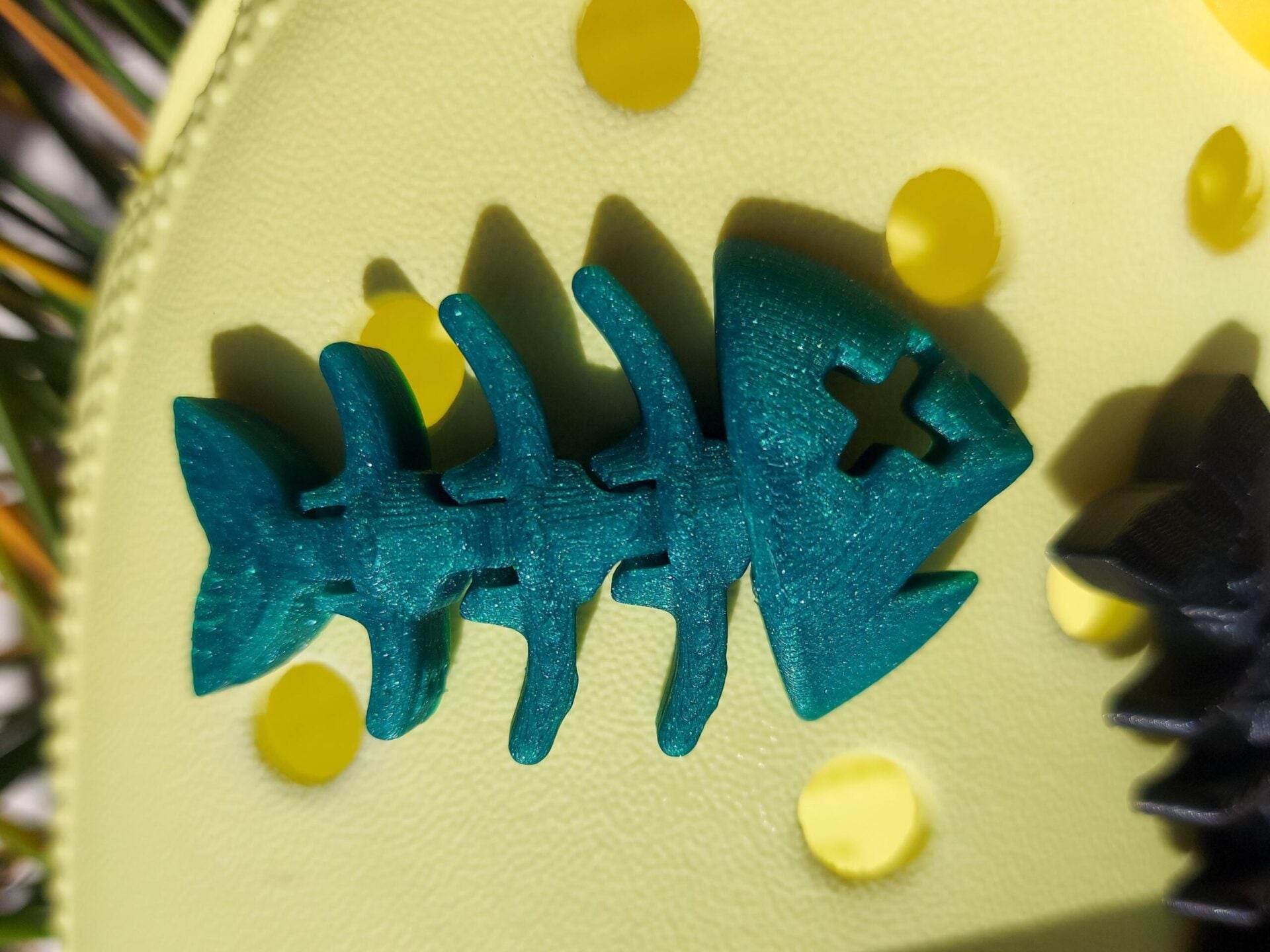 Yellow shoes adorned with colorful 3D-printed articulated fish bone charms in white, pink, black, teal, and yellow, displayed on a grassy background.