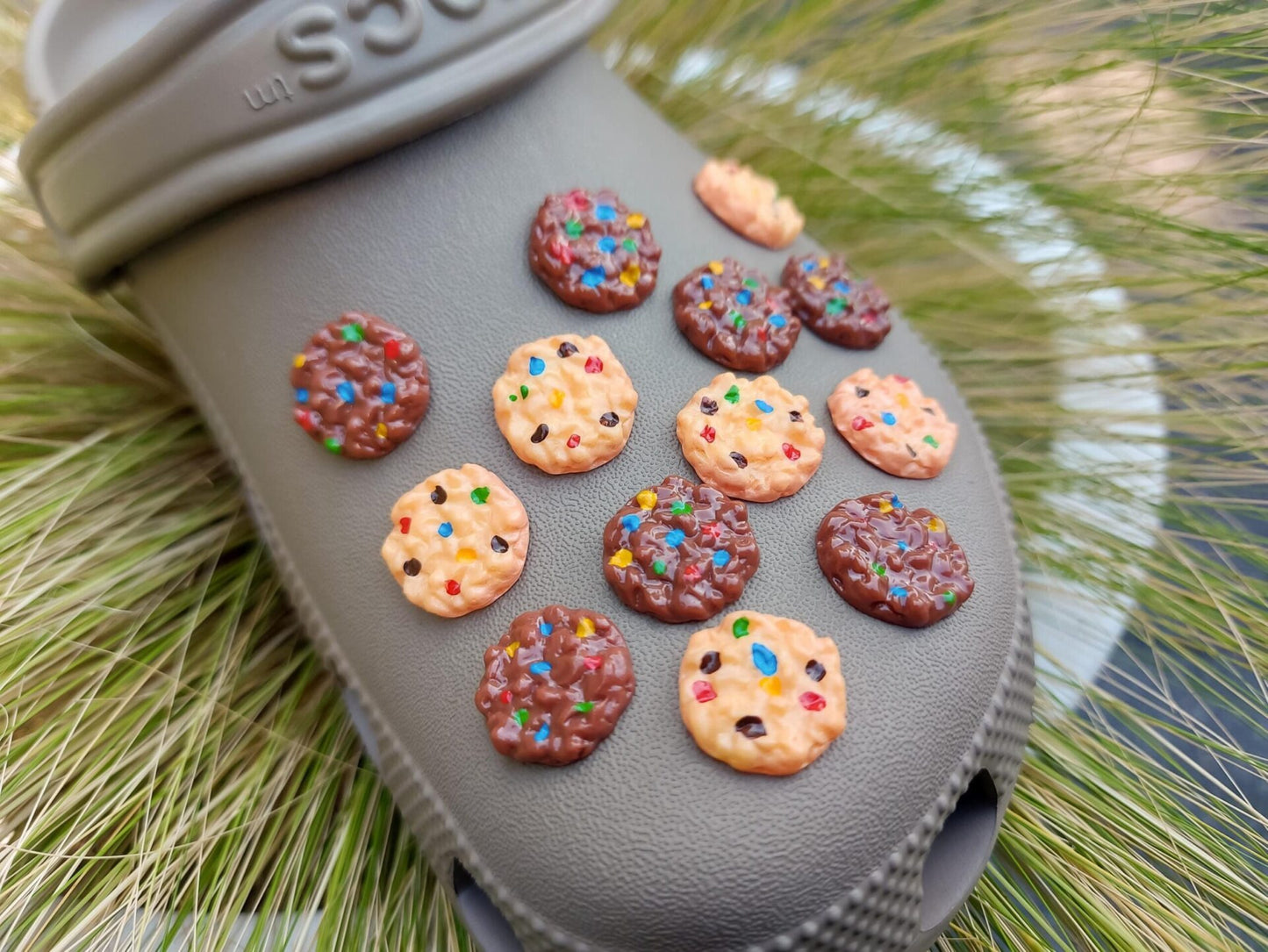 Kaki shoes decorated with various cookie charms, featuring chocolate chip and candy-topped cookies, displayed on a backdrop of green grass.