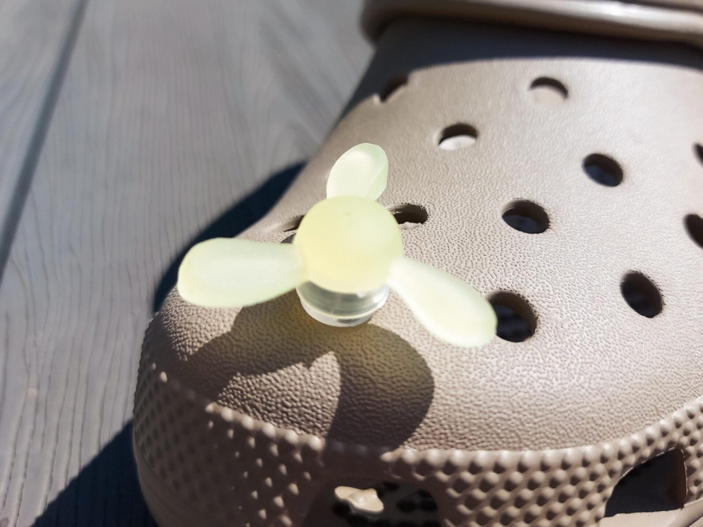 Close-up of glow-in-the-dark windmill shoe charms attached to a beige shoe, displayed outdoors in sunlight. The colorful windmill blades spin as you walk, adding a fun, dynamic feature to your shoes.