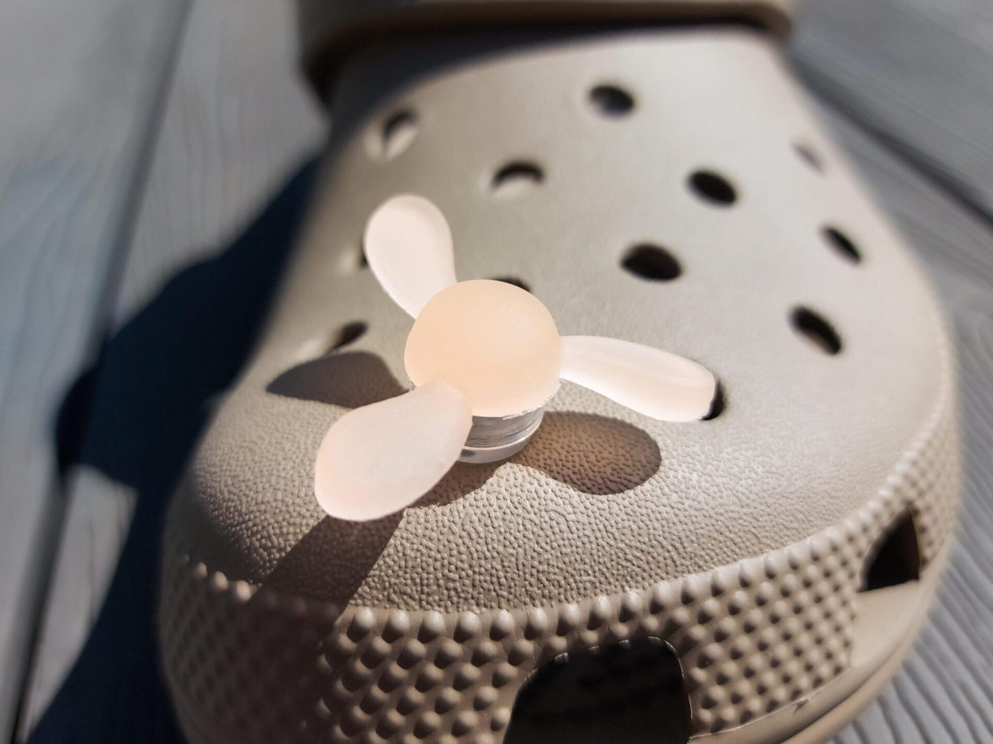 Close-up of glow-in-the-dark windmill shoe charms attached to a beige shoe, displayed outdoors in sunlight. The colorful windmill blades spin as you walk, adding a fun, dynamic feature to your shoes.