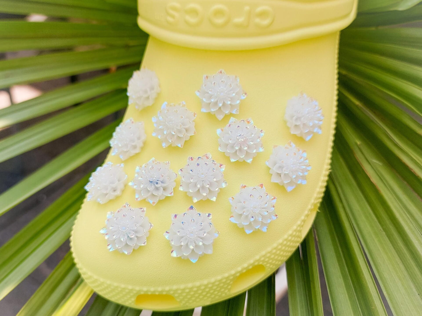 Yellow shoes adorned with multiple white rose charms featuring iridescent accents, placed on a background of green palm leaves. The charms add an elegant and sophisticated touch to the shoes.