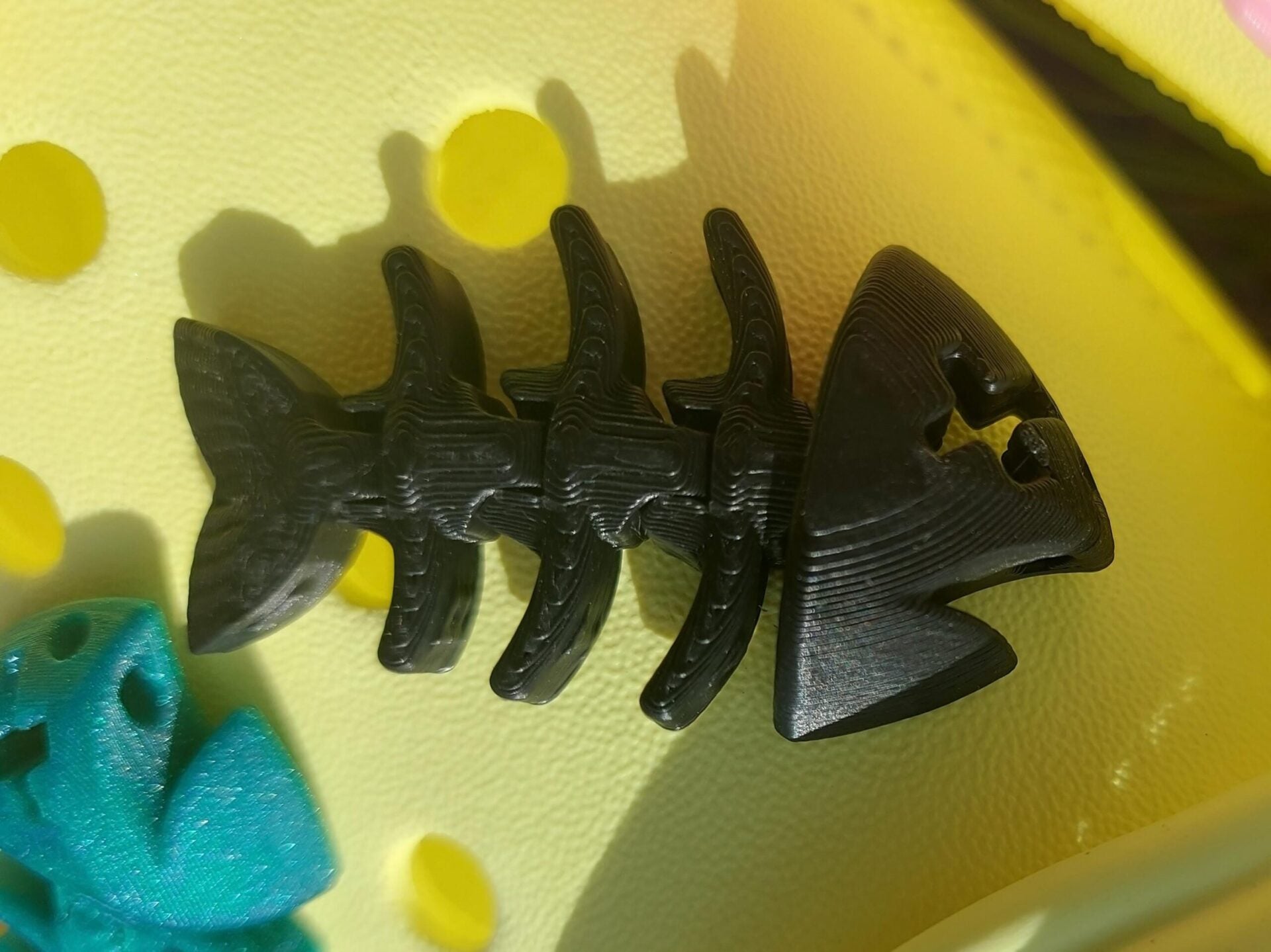 Yellow shoes adorned with colorful 3D-printed articulated fish bone charms in white, pink, black, teal, and yellow, displayed on a grassy background.