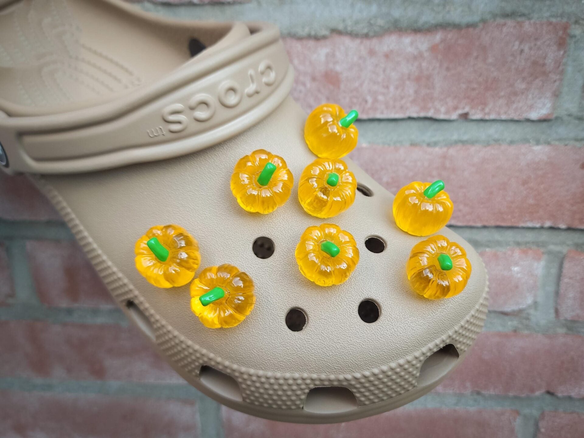 Kaki shoes adorned with small, glow-in-the-dark pumpkin charms, displayed against a brick wall background.