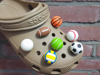 Close-up of a set of 7 sports-themed charms on a shoe shoe, featuring a basketball, football, soccer ball, tennis ball, baseball, volleyball, and golf ball. The charms are securely attached, adding a fun, athletic touch to the shoe.