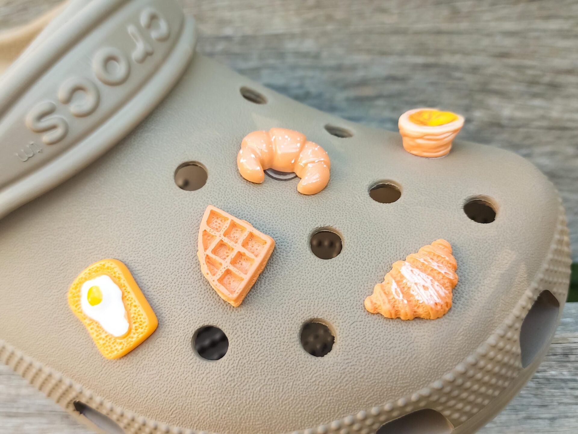 A playful assortment of breakfast-themed shoe charms featuring five distinct designs: two croissants (one classic and one chocolate), a golden pastry, a sunny-side-up egg on toast, and a waffle.