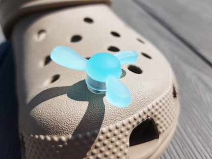 Close-up of glow-in-the-dark windmill shoe charms attached to a beige shoe, displayed outdoors in sunlight. The colorful windmill blades spin as you walk, adding a fun, dynamic feature to your shoes.