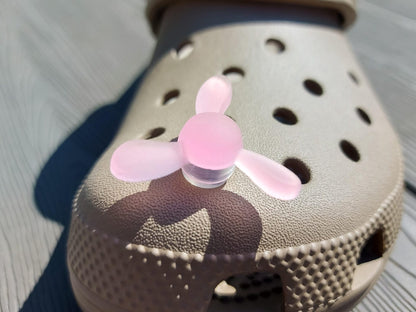 Close-up of glow-in-the-dark windmill shoe charms attached to a beige shoe, displayed outdoors in sunlight. The colorful windmill blades spin as you walk, adding a fun, dynamic feature to your shoes.