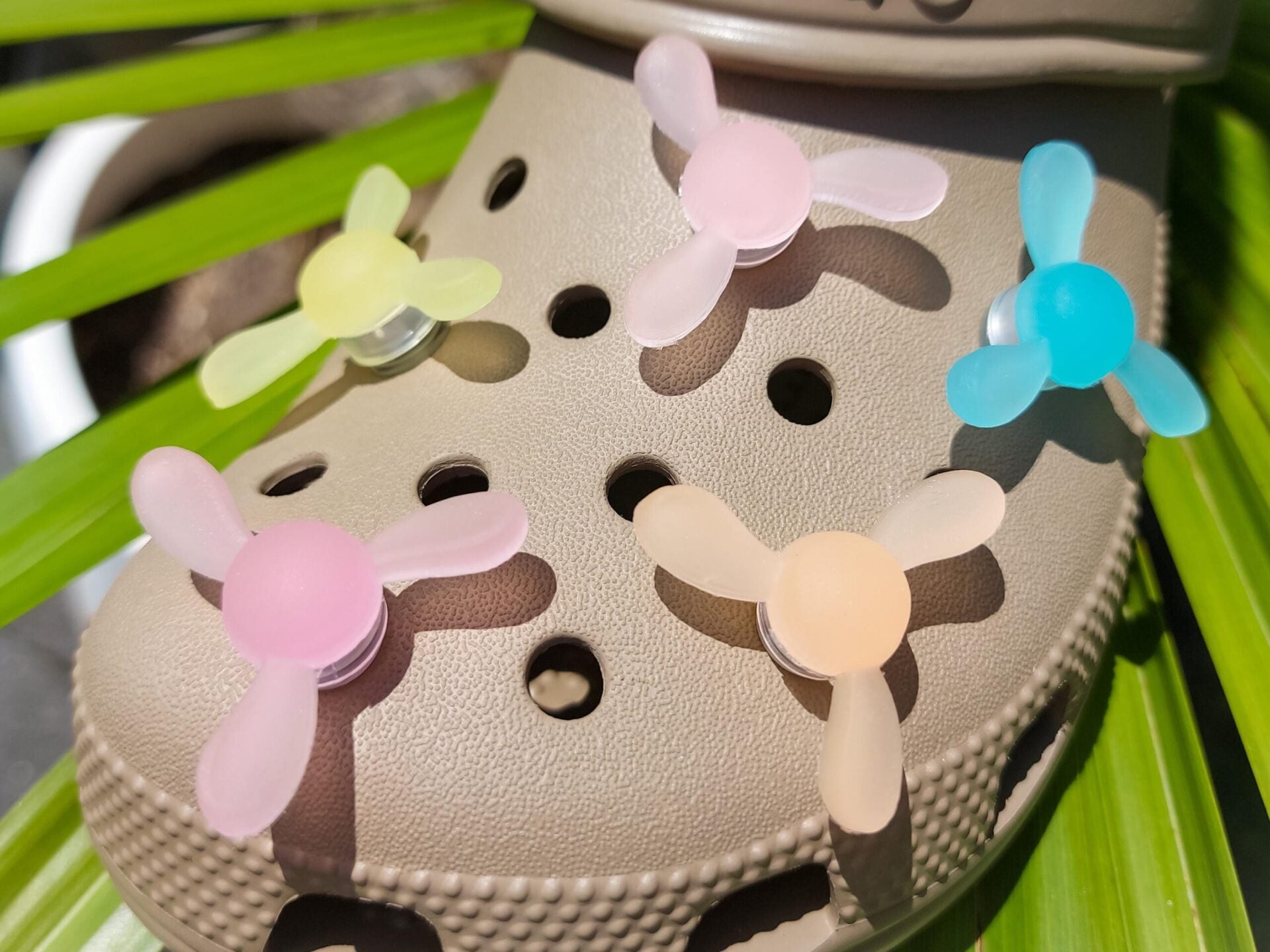 Close-up of glow-in-the-dark windmill shoe charms attached to a beige shoe, displayed outdoors in sunlight. The colorful windmill blades spin as you walk, adding a fun, dynamic feature to your shoes.