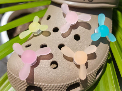 Close-up of glow-in-the-dark windmill shoe charms attached to a beige shoe, displayed outdoors in sunlight. The colorful windmill blades spin as you walk, adding a fun, dynamic feature to your shoes.